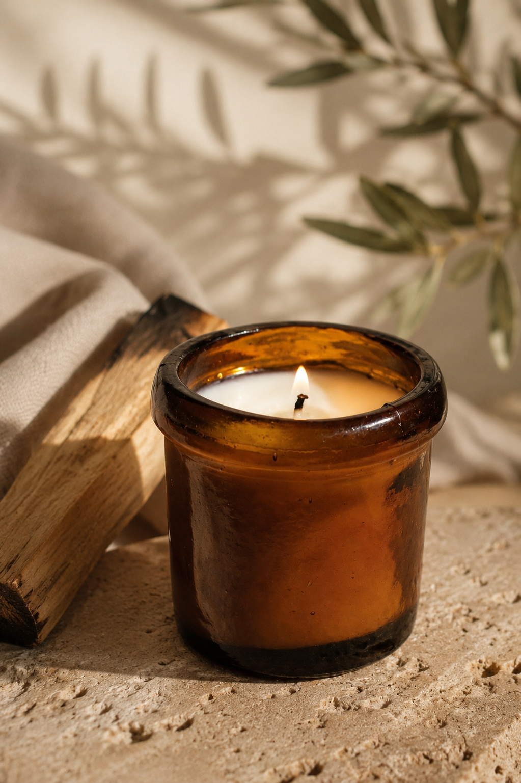 An amber glass jar candle with a lit wick sits on a textured stone surface next to a piece of charred wood, with soft shadows and olive branches in the background