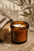 Load image into Gallery viewer, An amber glass jar candle with a lit wick sits on a textured stone surface next to a piece of charred wood, with soft shadows and olive branches in the background 
